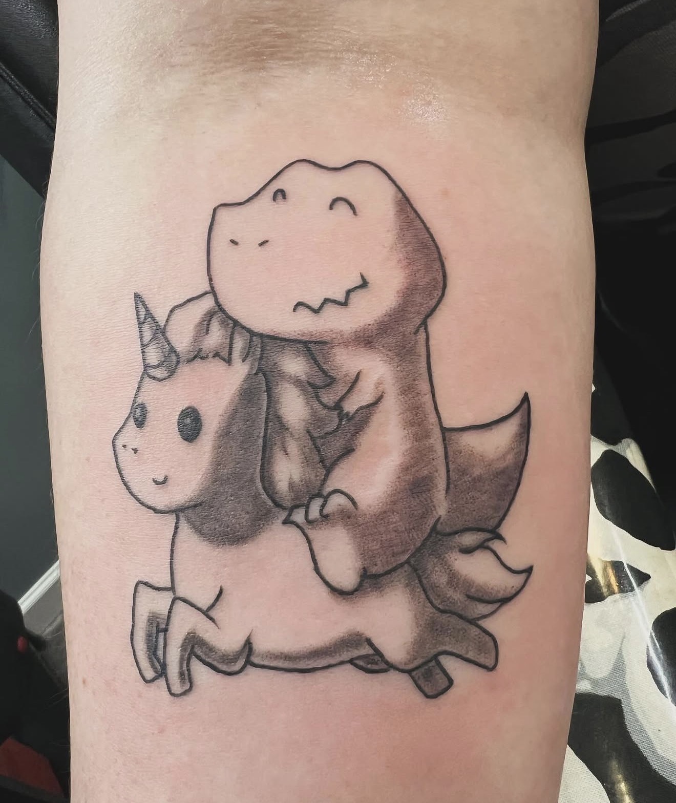 Cute black and grey dinosaur riding a unicorn forearm tattoo
